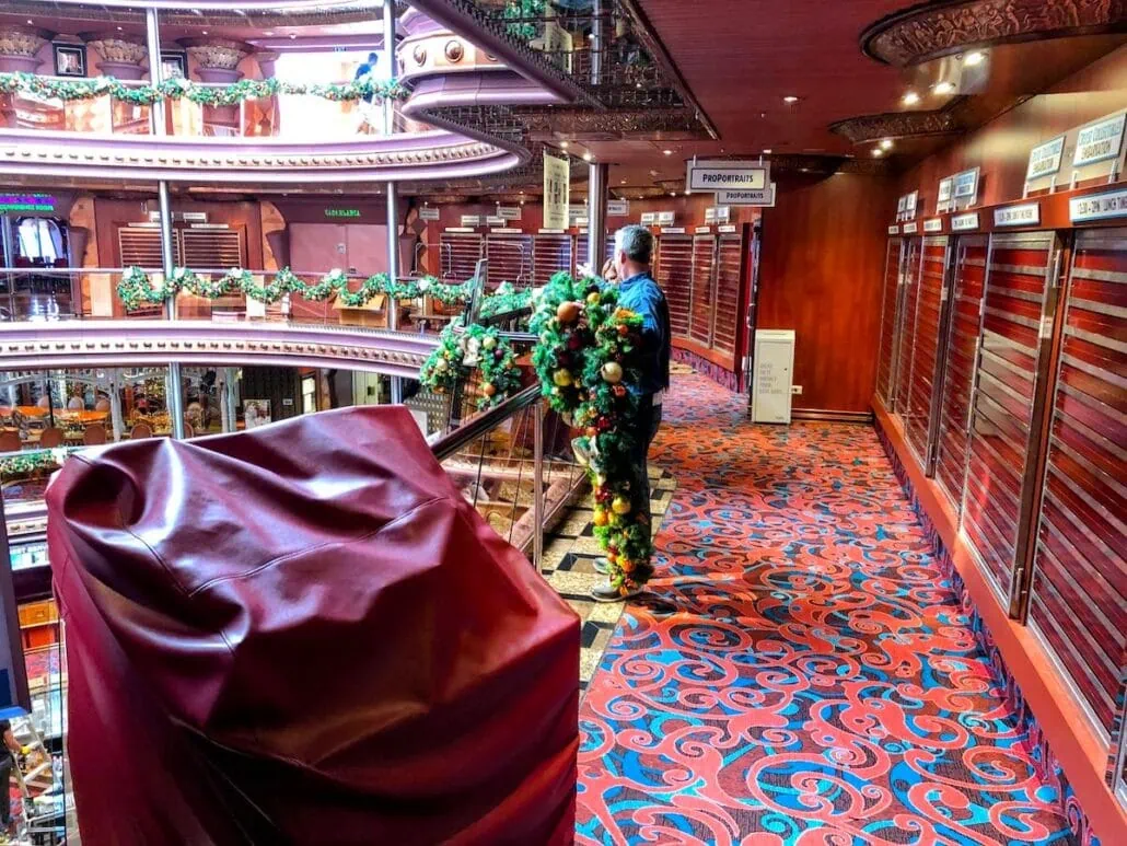 A person stands next to a railing in a decorated Carnival Cruise Line interior. The hallway features vibrant red and blue carpeting, rows of closed store shutters, and festive greenery with ornaments along the railing, showcasing a stunning holiday transformation.
