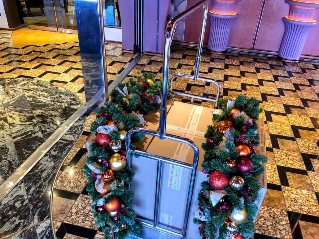 A luggage cart in the hotel lobby transforms into a festive spectacle, adorned with holiday wreaths featuring red and gold ornaments and lush greenery. The floor boasts a geometric black and gold pattern, while large pillars stand elegantly by the glass door, reminiscent of a Carnival Cruise Line ambiance.
