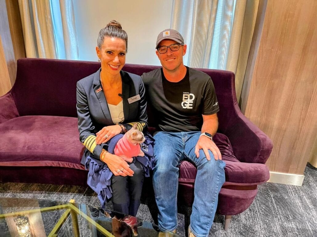 A woman in a uniform with name tags sits on a purple couch, holding a small dog dressed in pink, resembling the famed Popular Cruise Cat. Beside her, a man in an "EDGE" t-shirt smiles warmly. They are in a modern indoor setting, reminiscent of Celebrity Cruises' inviting interiors.