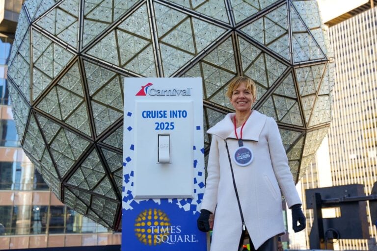 A person stands smiling in front of a large geodesic sphere in Times Square, with a sign reading "Cruise Into 2025" from Carnival. They are wearing a white coat and a round badge, reminiscent of an exciting TV commercial. Tall buildings form the backdrop.
