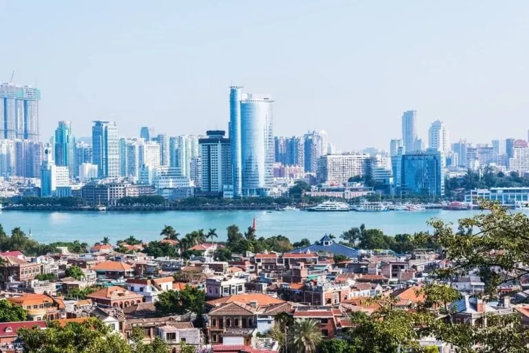 A cityscape in China showcases a vibrant mix of modern skyscrapers and traditional buildings. Thrill-seekers will enjoy the red-roofed houses and lush greenery in the foreground, while a wide river divides older structures from taller contemporary marvels in the background.