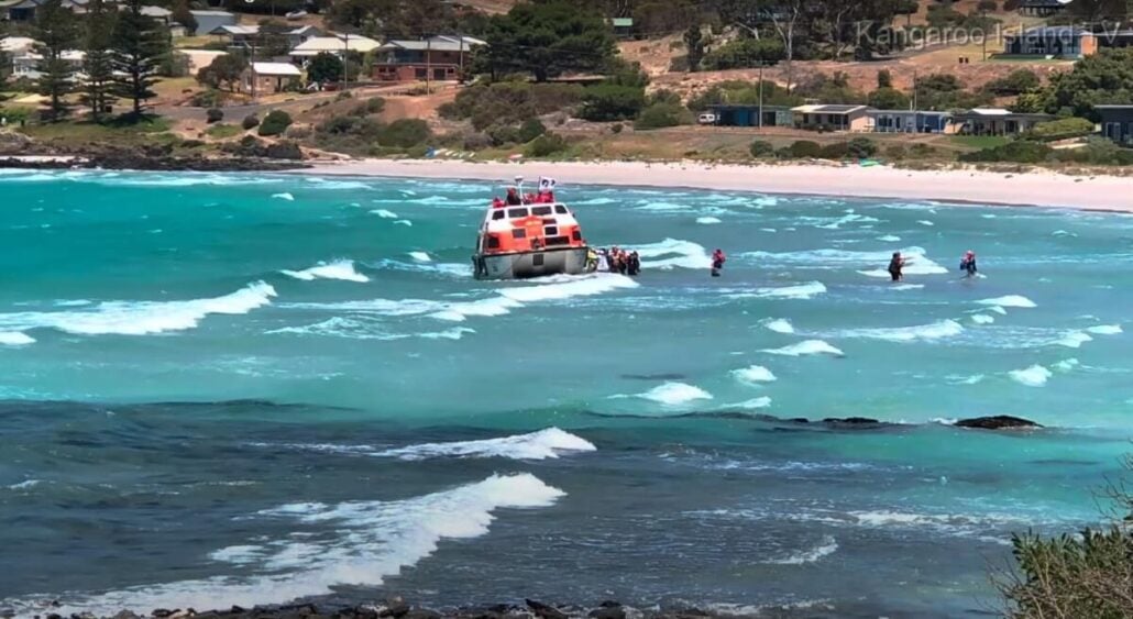 A rescue tender boat is being pushed into the turquoise ocean by several people in wetsuits. The scene is set against a backdrop of a sandy beach, trees, and houses on a hillside. Waves break gently around the boat, suggesting motion towards assisting passengers at sea.