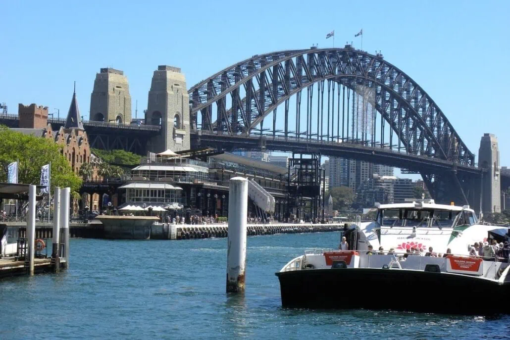 A sunny view of Sydney Harbour with the iconic Sydney Harbour Bridge in the background. The calm blue waters, dotted with docked boats, reflect a vibrant waterfront lined with buildings and trees. This scenic spot highlights a record-breaking season for the Australian cruise industry.