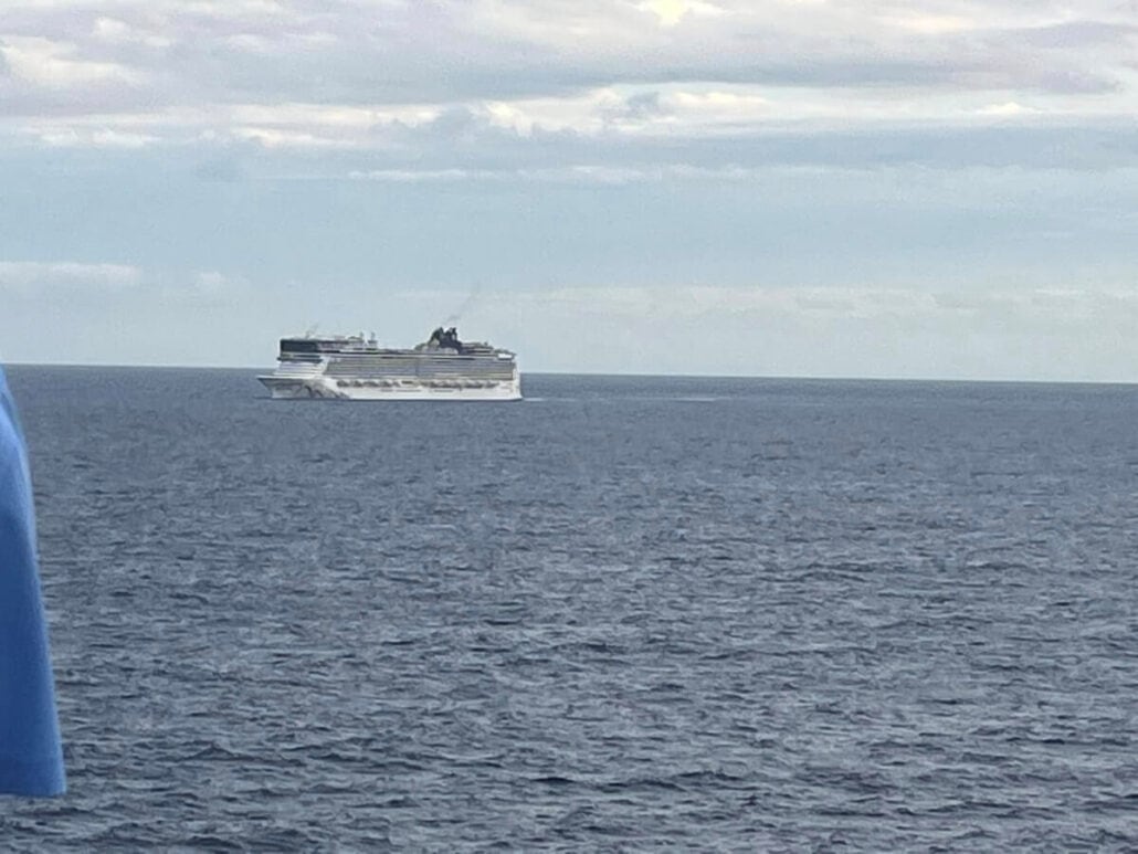 A Norwegian Cruise Line ship sails on a calm sea under a cloudy sky. In the foreground, part of a blue shirt hints at an overboard passenger story yet untold.