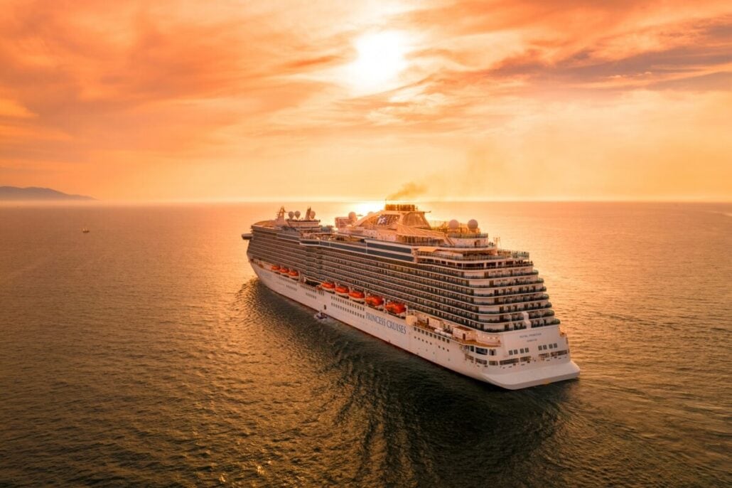 A large cruise ship with a bustling casino sails on calm waters during sunset. The sky, filled with orange and pink hues, reflects on the sea. The white ship boasts multiple decks, where gamblers enjoy entertainment. Lifeboats are visible on the side, and land is faintly visible on the horizon.