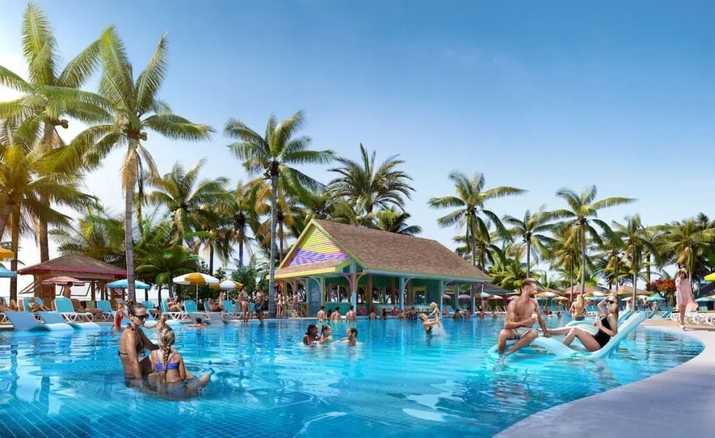 A lively swimming pool scene at the Royal Beach Club in Nassau, where adults and children enjoy the water. The pool, surrounded by palm trees and sun loungers, is set against a colorful building under the clear blue Bahamian sky.