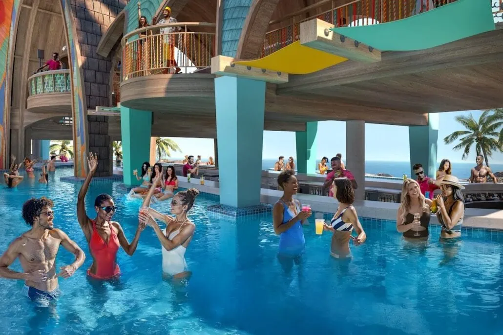 A lively pool scene unfolds at the Royal Beach Club, with people enjoying drinks and socializing at a swim-up bar. Some are in the pool, while others sit on submerged stools. With Nassau Bahamas' ocean and palm trees in the background, the setting is tropical and vibrant.