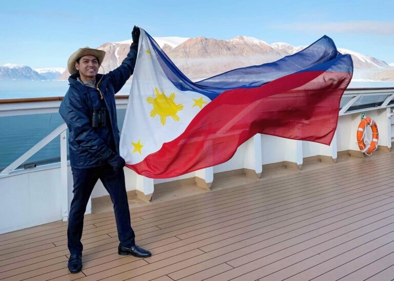 A person in a dark outfit and straw hat, resembling an influencer on their one day off, holds the Philippine flag on the deck of a ship. The backdrop reveals a stunning contrast of water and snow-covered mountains beneath a clear sky.