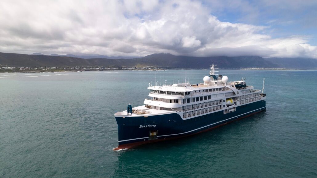 A large blue and white cruise ship, named SH Diana, sails on calm waters. The sky is partly cloudy, and distant mountains can be seen on the horizon. Despite the canceled Antarctica visit, irate passengers remain hopeful for new adventures.