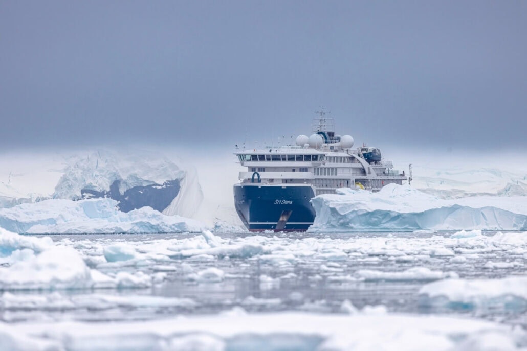 A cruise ship navigates through icy waters surrounded by icebergs and snow-covered terrain under a cloudy sky. The irate passengers aboard express their frustration over the canceled Antarctica visit, feeling the disappointment of a missed adventure in this polar region.