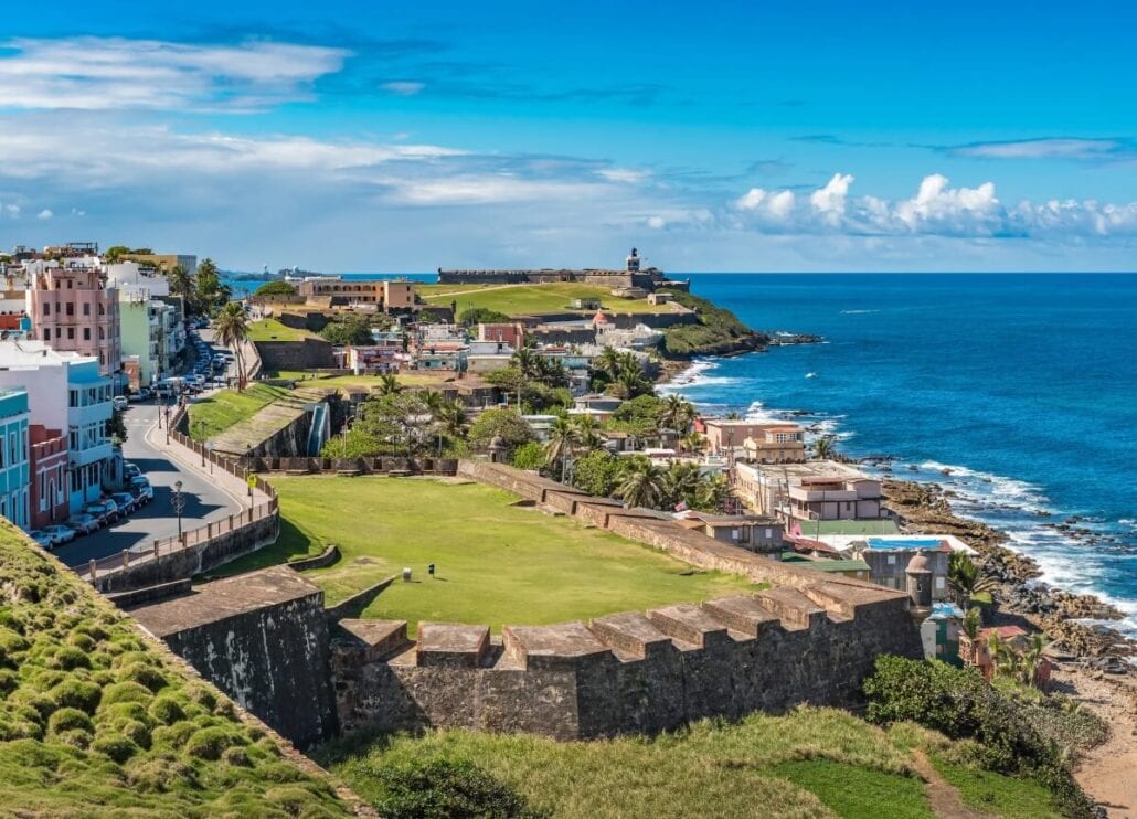 A scenic view of San Juan, Puerto Rico, featuring the historic fortress of El Morro and vibrant buildings along the coastline. The image captures lush greenery and the blue ocean under a clear sky, highlighting popular cruise itineraries that often include a stop at San Juan Pier.