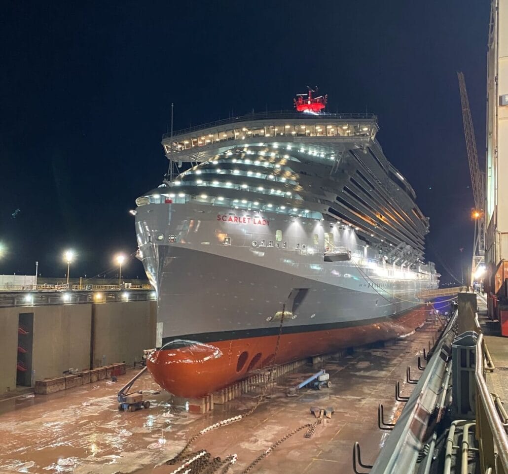 A large cruise ship, the Scarlet Lady, is docked at night in a shipyard, illuminated by bright lights. The vessel gleams red and white, hinting at recent upgrades during its dry dock.