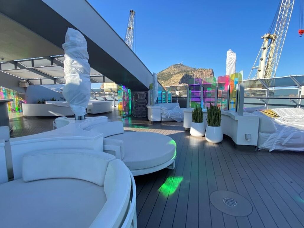 A modern outdoor lounge area on the Scarlet Lady yacht deck features rounded white seating, potted plants, and colorful glass panels. Wrapped structures are visible, hinting at recent upgrades. Cranes and a mountain are in the background under a clear blue sky.