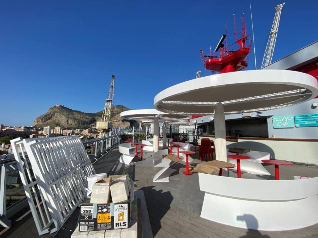 The outdoor seating area on the Scarlet Lady's deck features round tables and red chairs under large white canopies. Amid boxes and construction materials for dry dock upgrades, a port, cranes, and mountains are visible in the background under a clear blue sky as the ship prepares to return to service.