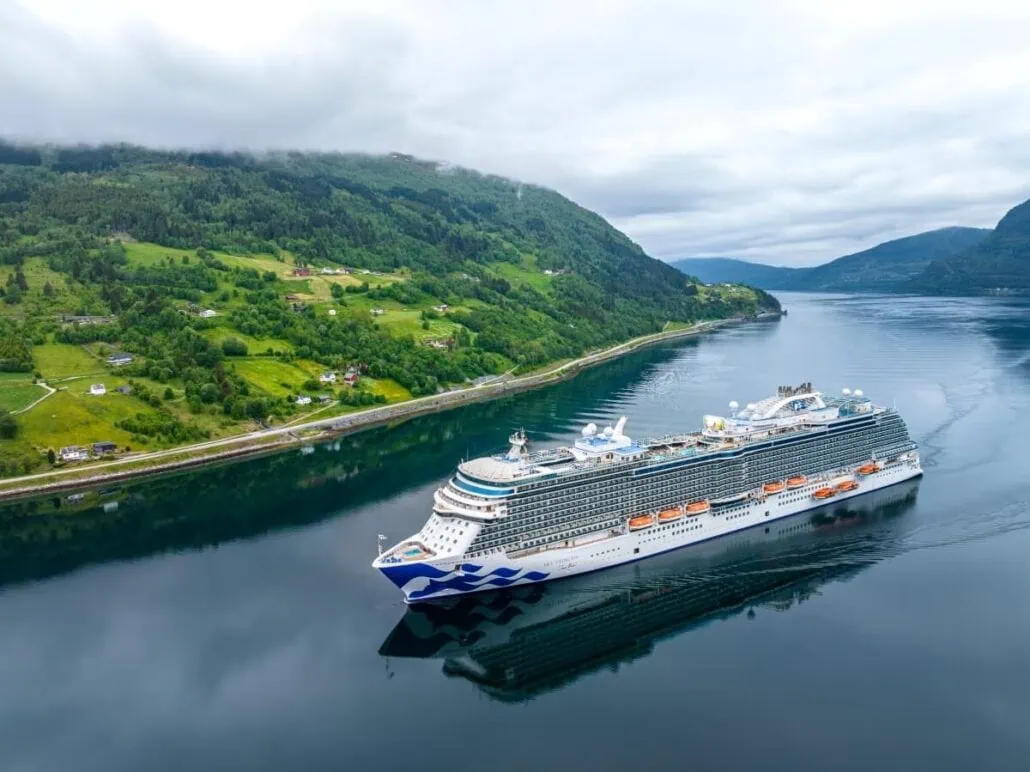 A large Princess Cruises vessel sails through a calm fjord, surrounded by green hills and scattered houses. The sky is overcast, reflecting in the still waters. The ship is near the coastline, highlighting the serene, picturesque landscape.
