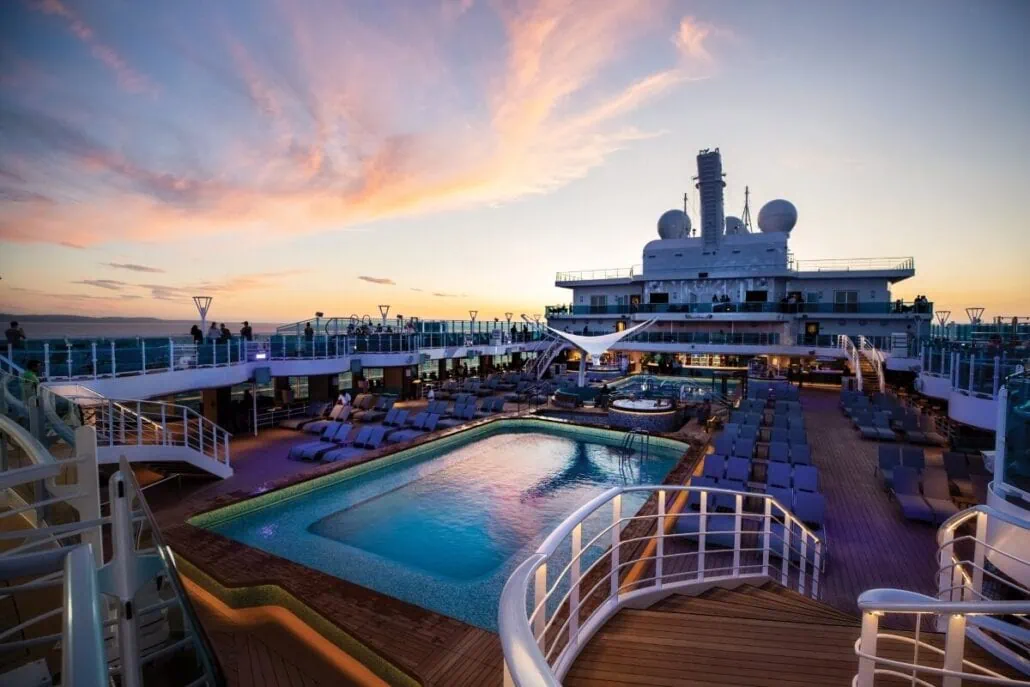 A luxurious Princess Cruises deck at sunset features a central pool surrounded by lounge chairs. Soft lighting illuminates the area as the sky displays pink and orange hues. Antennas and radar domes are visible, with an elderly female enjoying the serene evening air.