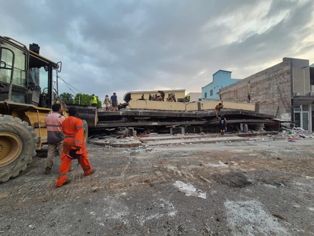 Rescue workers in orange and yellow uniforms navigate the rubble around a collapsed building, showcasing the dedicated spirit of Vanuatu Earthquake Relief Efforts. A large construction vehicle is on the left under an overcast sky, symbolizing hope amidst adversity.