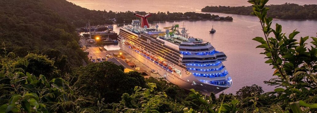 A large cruise ship, part of a Carnival fleet, docked at a port surrounded by lush green hills. The ship is illuminated with blue lights and the sun is setting in the background, casting a warm glow over the water and landscape.