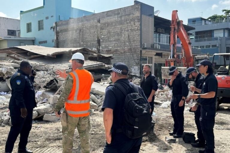 A group of people, including a person in an orange vest and a white hard hat, and several in dark uniforms, stand in front of a pile of rubble from the Vanuatu Earthquake. An excavator is in the background as part of the relief efforts. Buildings surround the area on a bright day.