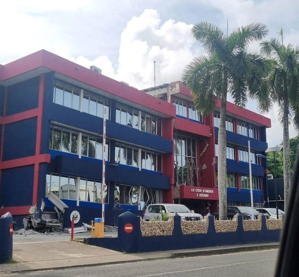 A three-story building with a blue and red facade shows signs of damage, likely from the Vanuatu Earthquake, with debris scattered around. Palm trees stand nearby, and several parked cars are in front. A sign on the building reads "La Casa de la Cultura Ecuatoriana.