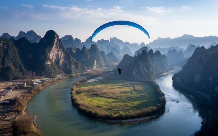 A paraglider with a blue parachute soars over a lush, green landscape, enticing thrill-seekers with an adrenaline-pumping adventure activity. The winding river and surrounding mountains create a breathtaking view under the clear blue sky.