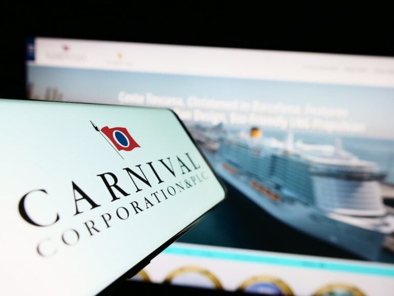 A smartphone screen shows the Carnival Corporation logo in front of a blurred computer screen displaying an image of a cruise ship and text.