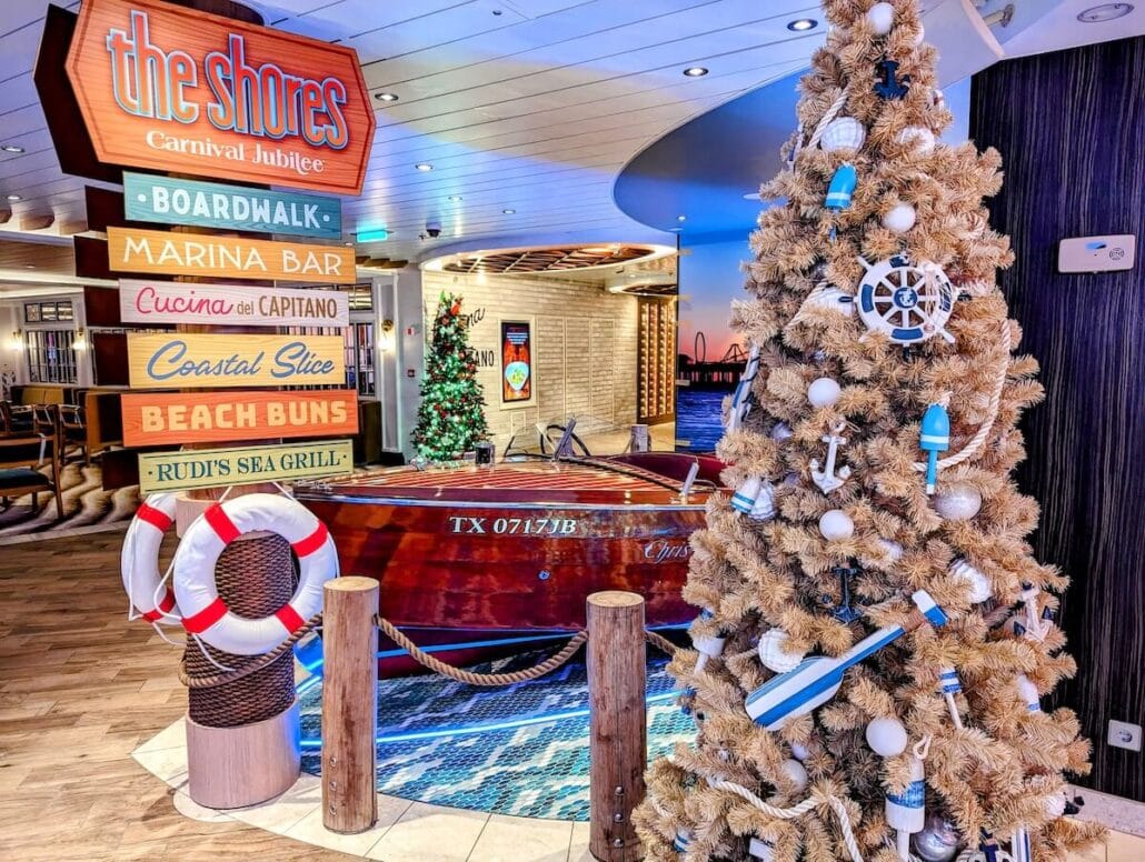 A festive Carnival Cruise Line interior, decorated for the holidays, features a beige Christmas tree adorned with blue and white ornaments. Nearby, a classic wooden speedboat and a colorful sign point to various holiday-themed ship attractions, like bars and restaurants.