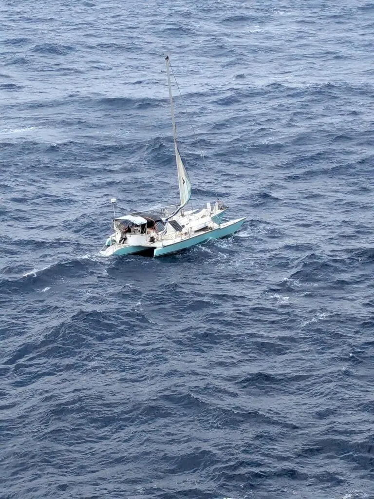 A small sailboat with a single mast navigates through choppy ocean waves, looking like it might belong to the fleet of Celebrity Cruises. The sea is dark blue and the sky is obscured as the vessel tilts slightly amidst the surrounding water.