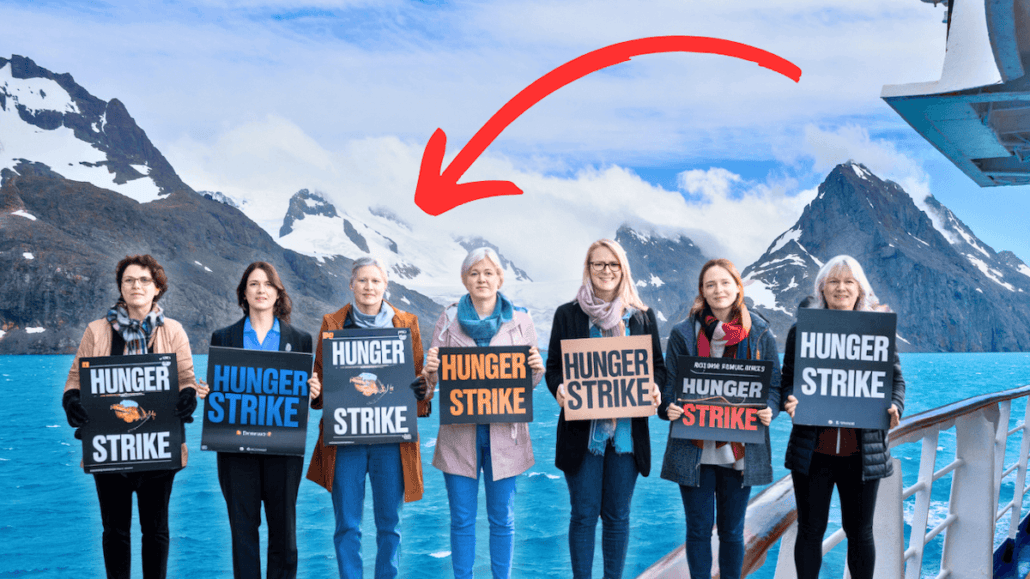 A group of seven irate passengers stands on a ship deck with snowy mountains and a blue ocean as the backdrop. They hold signs that read "Hunger Strike" during their Antarctica visit, with a red arrow pointing to the person in the center.