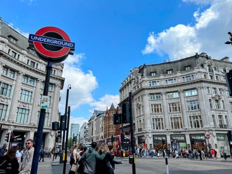 A busy urban intersection features an Underground sign in the foreground. Classic European architecture surrounds the street, filled with Visa-Exempt Travelers and a red double-decker bus under a partly cloudy sky.