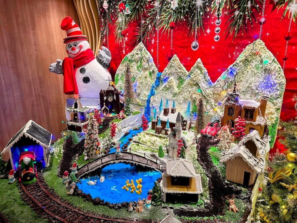 A festive holiday display features a snowman, mountains, a charming village with houses, a church, and a pond with ducks. A small train gracefully follows its tracks while lights and red ornaments sparkle against the greenery under a vibrant red backdrop. It's like capturing Carnival Cruise Line's transformative magic on land.