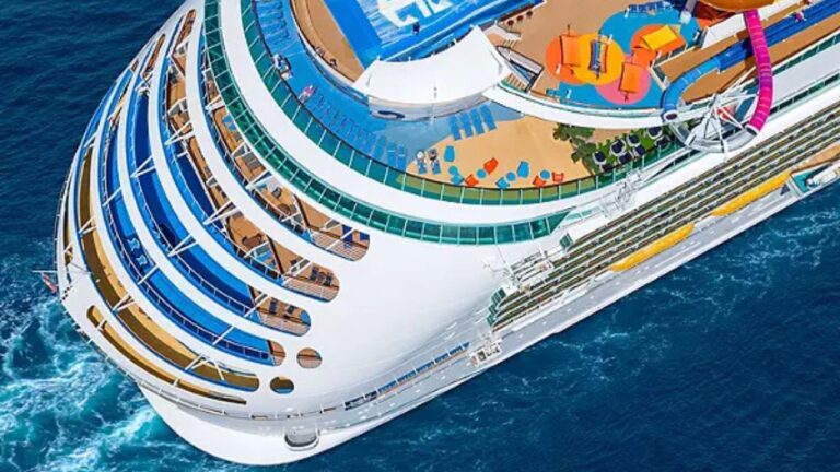 Aerial view of a Royal Caribbean cruise ship in the ocean. The ship boasts a white hull with blue and orange accents. Visible are multiple decks featuring a swimming pool, sun loungers, and colorful areas, where passengers enjoy amenities like water slides and a flowrider on the upper deck.