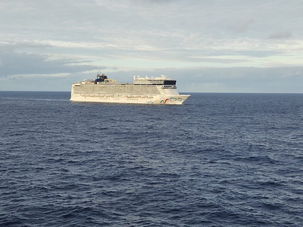 A large white Norwegian Cruise Line ship sails on a vast expanse of blue ocean under a cloudy sky. The vessel, decorated with colorful designs on its side, stands proudly as the horizon stretches in the background, its search ends with endless adventures awaiting on board.
