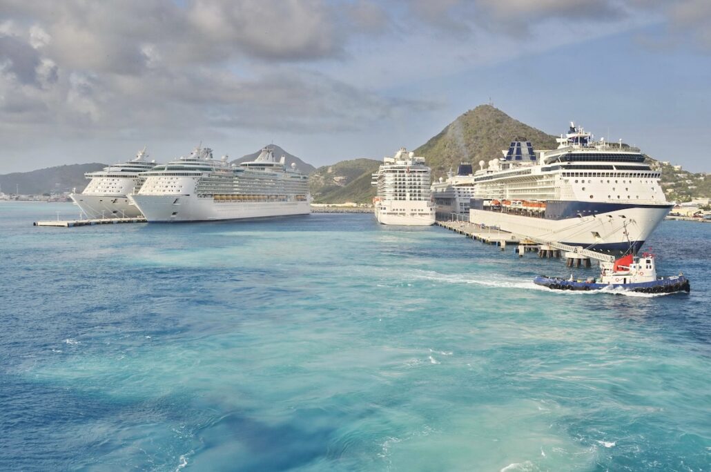 Four large cruise ships, possibly carrying excited passengers, are docked at a pier in St. Maarten's tropical harbor, with clear blue water in the foreground. A small tugboat navigates near the pier as lush green hills rise majestically under a partly cloudy sky.