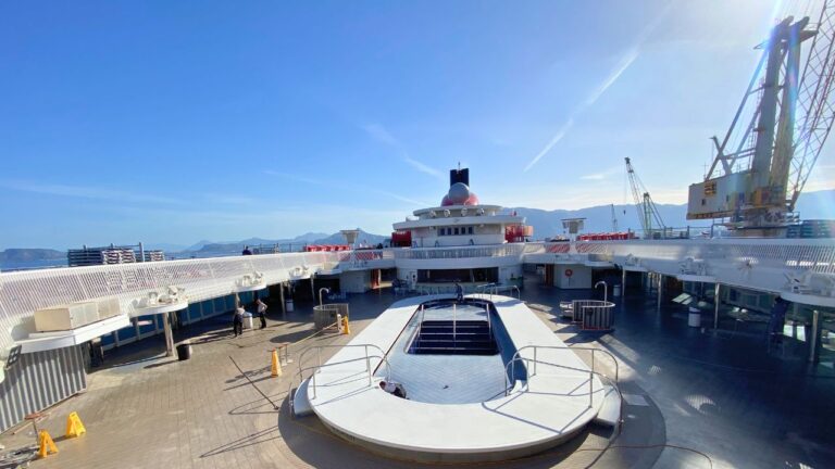 An empty cruise ship deck under a clear blue sky on the Scarlet Lady, featuring a covered pool at the center. Surrounding structures include lifeboats, cranes, and railings, hinting at recent dry dock upgrades. The background shows distant mountains and a bustling harbor area.