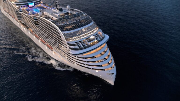A large MSC cruise ship sails on the open sea, viewed from above. The vessel features multiple decks, illuminated windows, and a swimming pool on the top deck. The water is dark and calm as the ship moves forward, creating a wake—a scene reminiscent of a Super Bowl commercial with surprise celebrities.