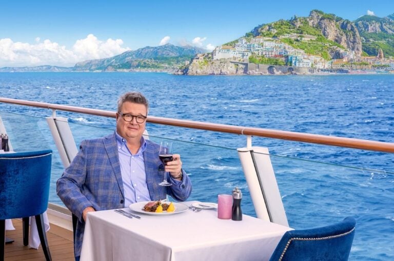 Eric Stonestreet, NCL’s Brand Ambassador and award-winning actor, sits at a dining table on a cruise ship deck, holding a glass of red wine. A meal is set before him. In the background, an ocean view and rocky, green coastal landscape with buildings are visible under a blue sky.