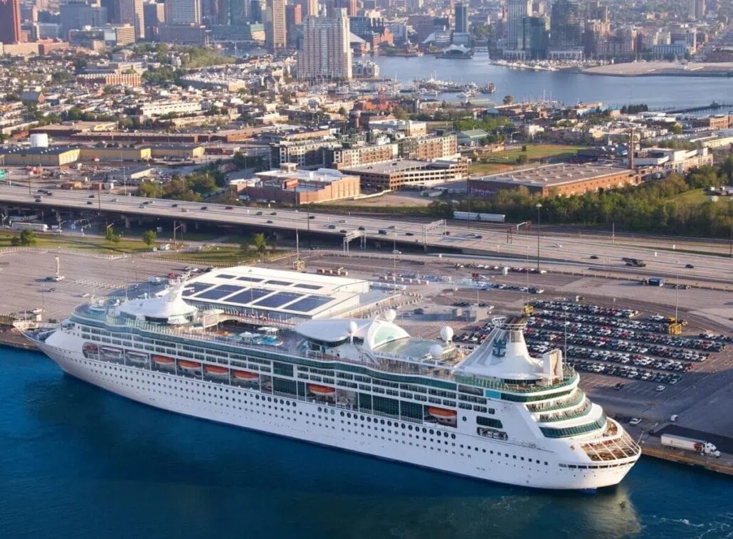 An aerial view captures a large white Royal Caribbean cruise ship docked at a port with a city skyline in the background. The vessel boasts multiple decks and a blue trim, with parked cars near the dock and buildings in the distance.