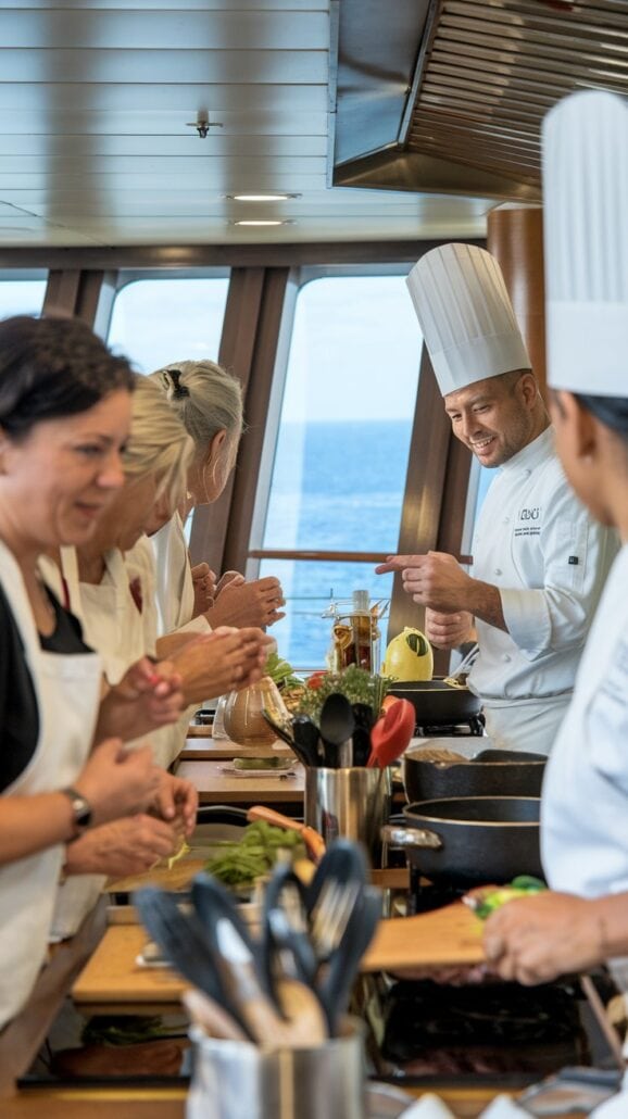 A group of people in a ship kitchen are being instructed by a chef in a white uniform and tall hat. They are preparing ingredients on wooden countertops, with pots, pans, and fresh produce visible. The ocean is seen through large windows in the background—a perfect gift idea for culinary enthusiasts on a cruise.