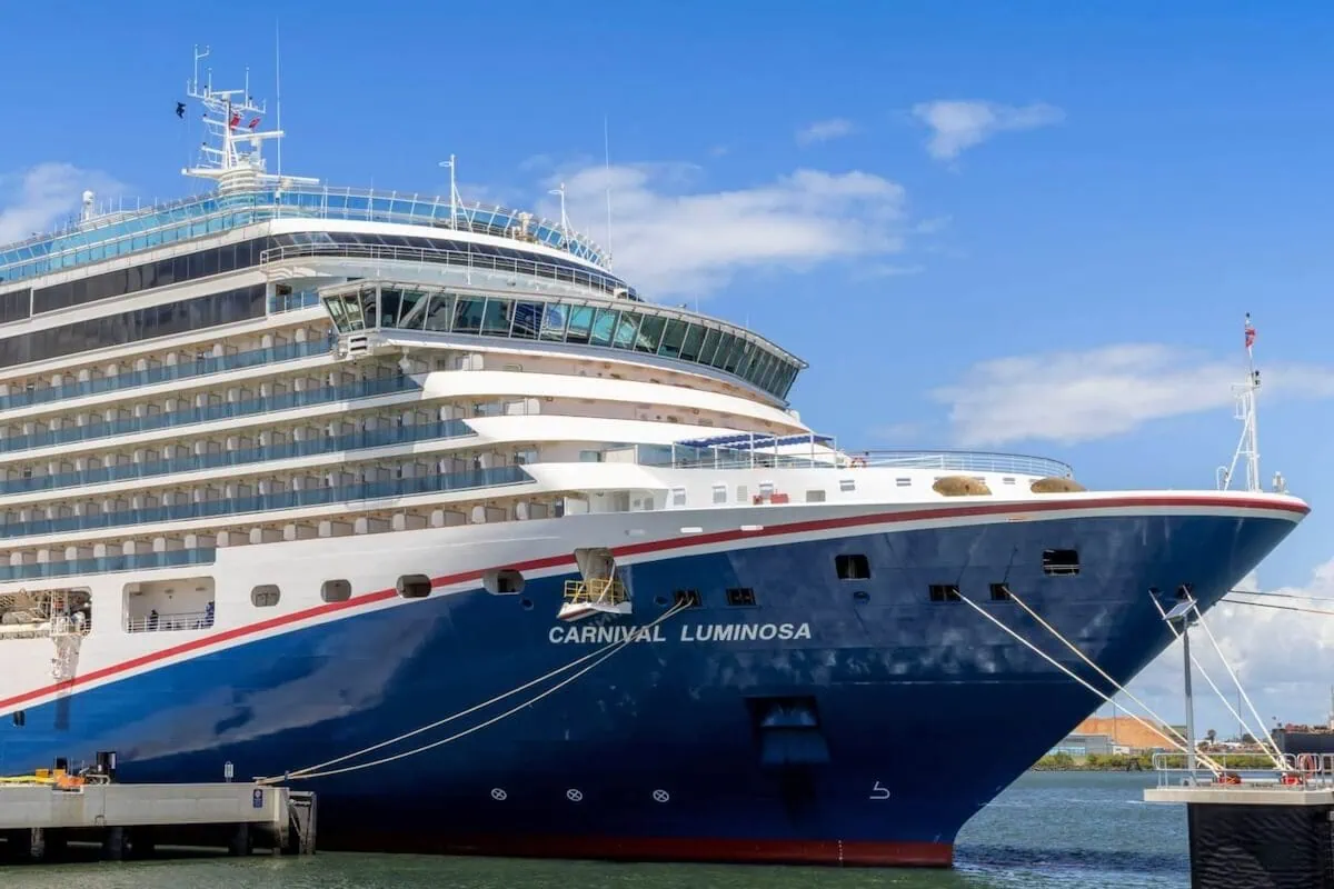 The Carnival Luminosa, known for its Asia-Pacific sailings, is docked at the port. The ship boasts a blue hull with a white superstructure and distinctive red and white striping, set against a clear sky with some clouds and calm waters.