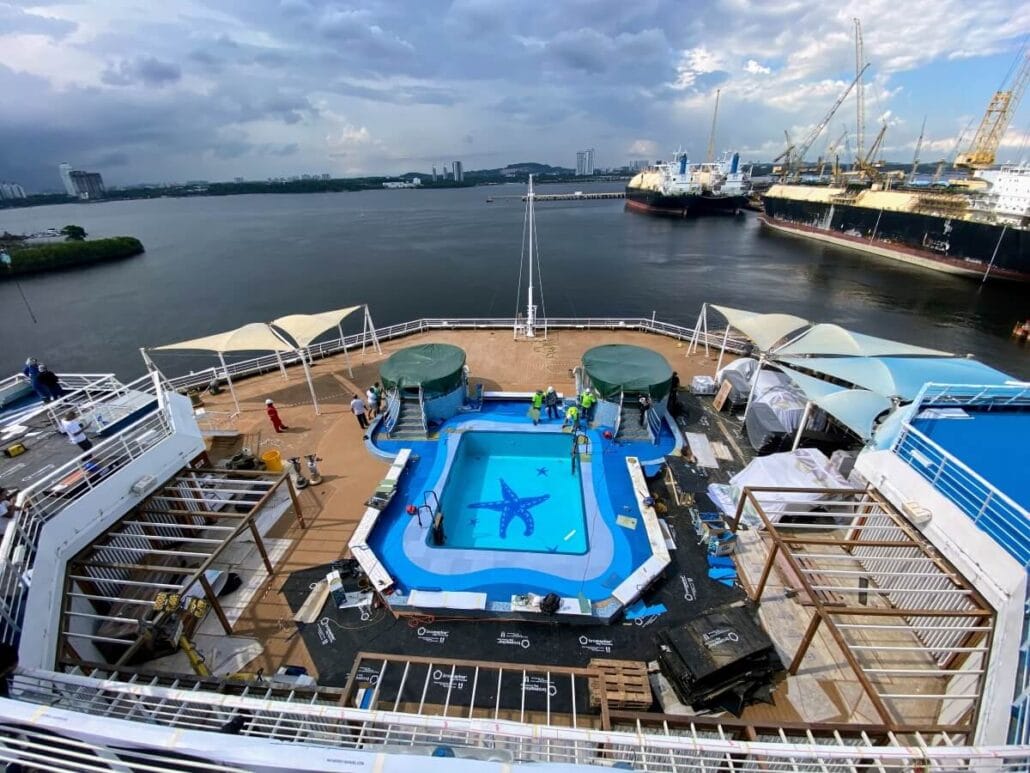 View from above of the Carnival Splendor's deck, highlighting a small swimming pool with a blue starfish design. Crew members stand nearby amidst various equipment, prepping for the 2024 dry dock changes. The ship rests by an industrial port under a cloudy sky.