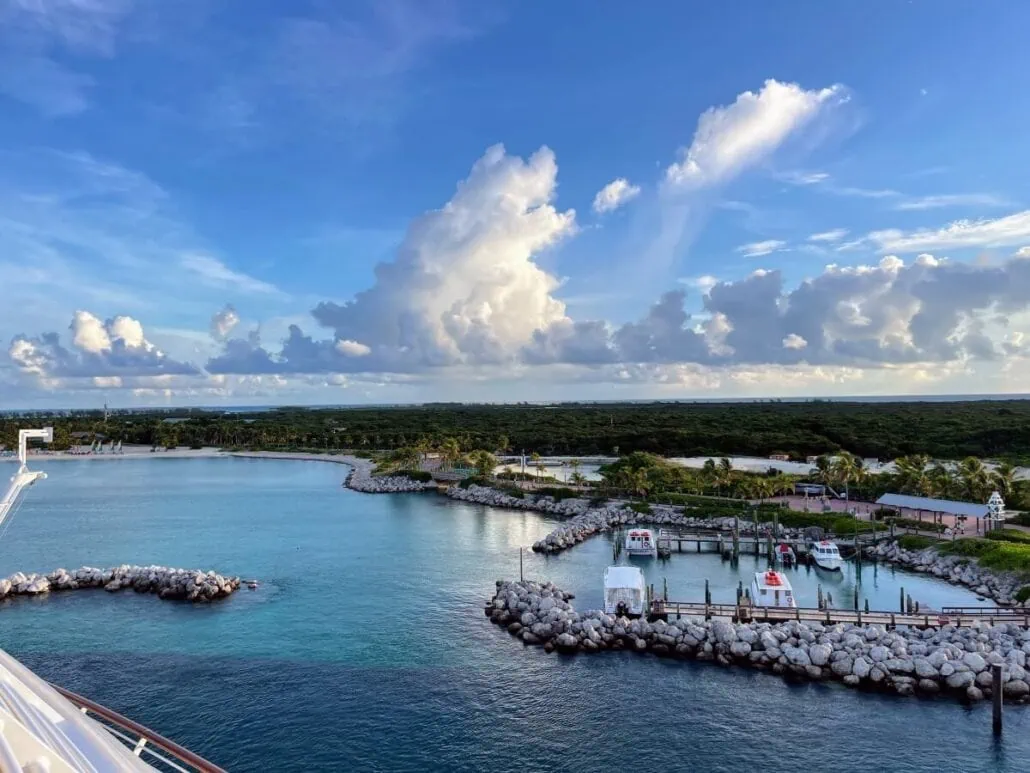 Nestled in a coastal area resembling Castaway Cay, a small marina cradles several boats, shielded by rocky barriers and lush greenery. The partly cloudy sky allows the sun to dance on the blue-green waters, creating a serene and picturesque setting.
