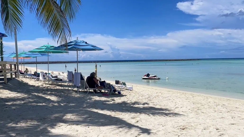 A sandy beach with lounge chairs and umbrellas at Castaway Cay is shown. People are relaxing under the umbrellas near the water. The ocean is calm and blue, with a small boat floating near the shore. Palm fronds hang overhead, and the sky is partly cloudy.