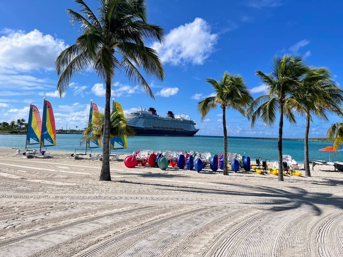 A tropical beach on Castaway Cay boasts several palm trees, colorful sailboats, and pedal boats on the sand. A large cruise ship is docked in the turquoise water in the background, while the sky remains mostly clear with a few clouds.