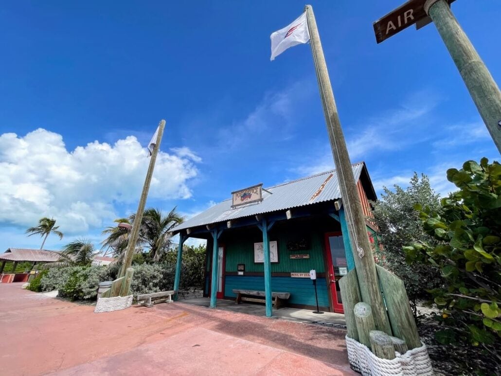 Under the bright blue sky of Castaway Cay, a small wooden building with a corrugated metal roof and teal walls stands. Two tall wooden poles with white flags flank it, surrounded by palm trees and bushes. A sign reads "Air" with an arrow, blending rustic charm with island vibes.