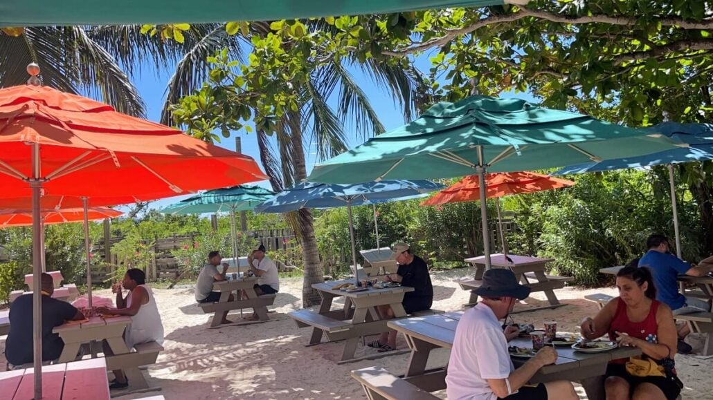 People sit at picnic tables under colorful umbrellas, including red, orange, and turquoise, set on a sandy area with tropical plants and trees around. The atmosphere is relaxed and sunny, evoking the charm of Castaway Cay’s outdoor cafes or beachside dining spots.