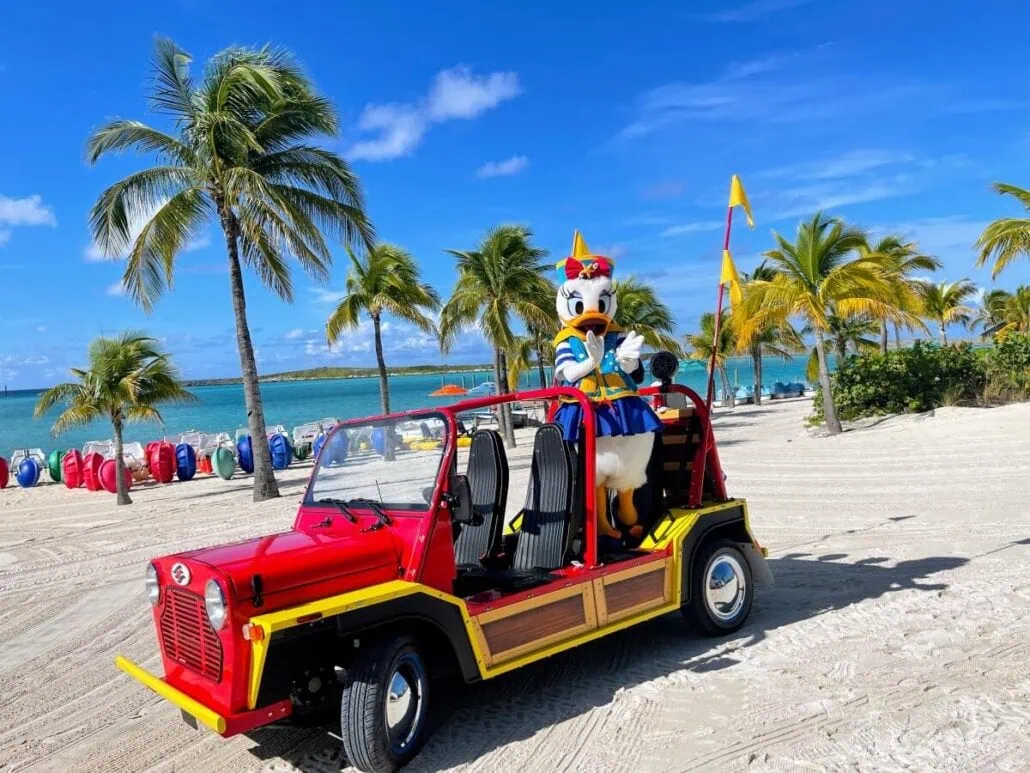 A costumed character resembling a duck sits in a red open-top vehicle on the sandy shores of Castaway Cay. Palm trees, colorful buoys, and turquoise waters shimmer under a blue sky with a few scattered clouds.