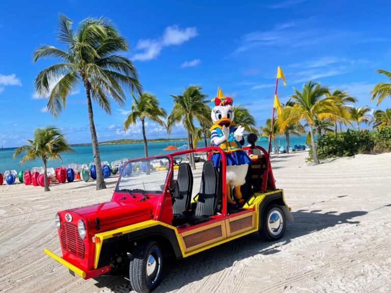 A costumed character resembling a duck sits in a red open-top vehicle on the sandy shores of Castaway Cay. Palm trees, colorful buoys, and turquoise waters shimmer under a blue sky with a few scattered clouds.