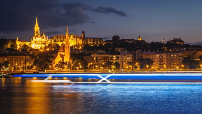 A nighttime view of a cityscape with illuminated historic buildings, including a tall church, along a riverbank. Bright blue and white streaks from a passing river cruising boat are visible on the water in the foreground.