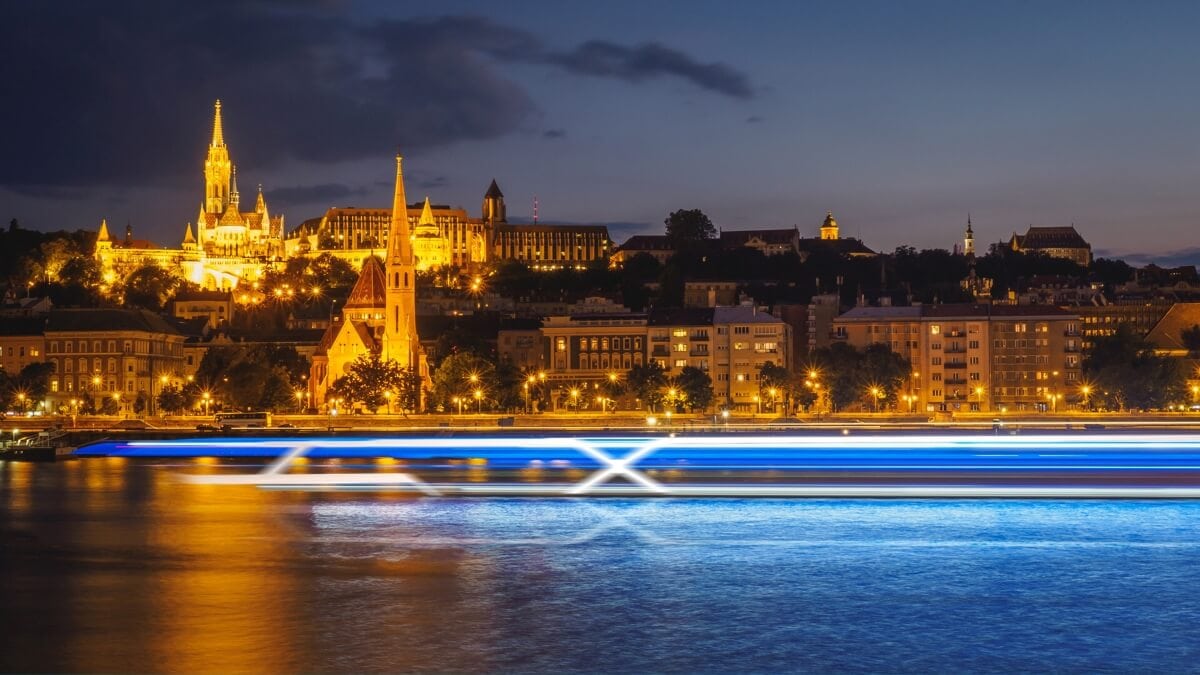 A nighttime view of a cityscape with illuminated historic buildings, including a tall church, along a riverbank. Bright blue and white streaks from a passing river cruising boat are visible on the water in the foreground.